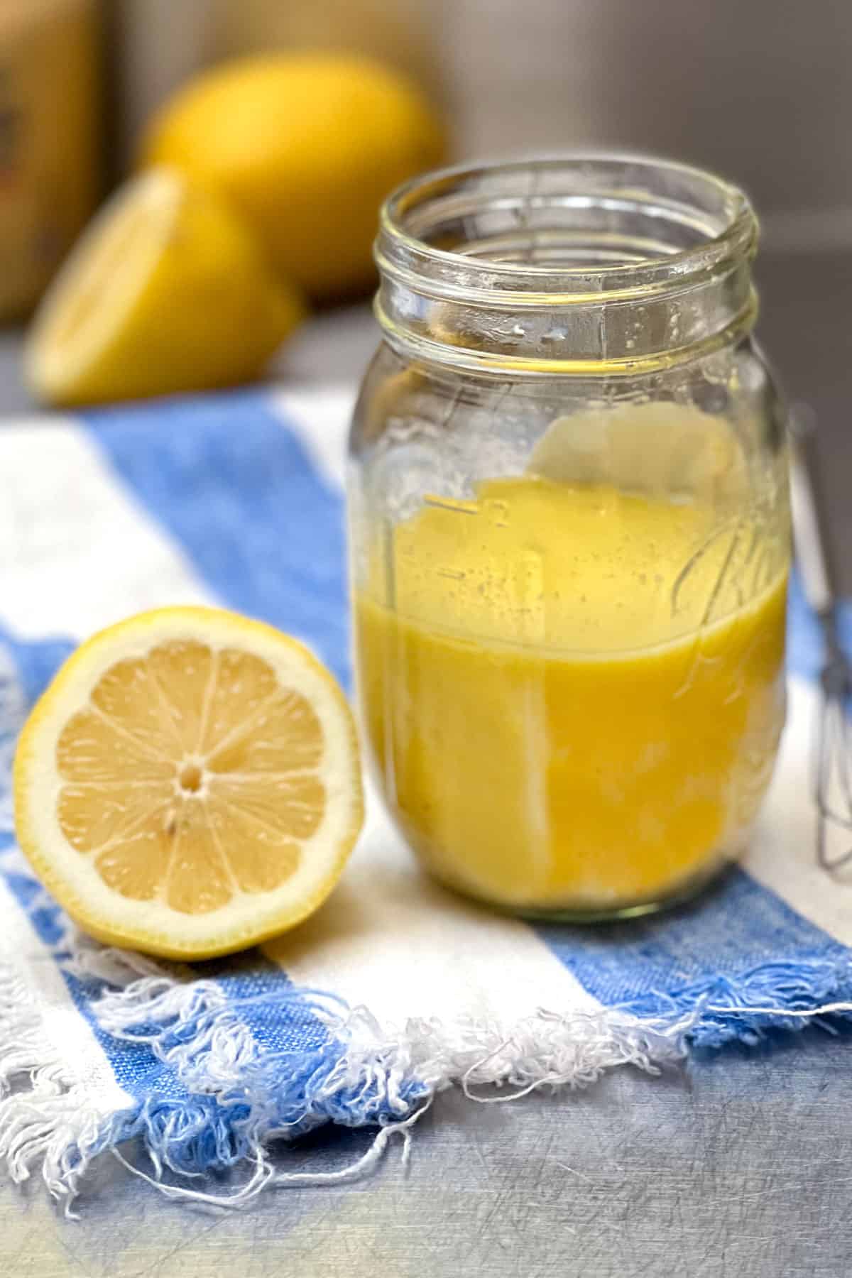 A mason jar on a striped cloth napkin, filled halfway with lemon vinaigrette, half a sliced lemon sitting next to it and a few lemons in the background, small wire whisk to the side