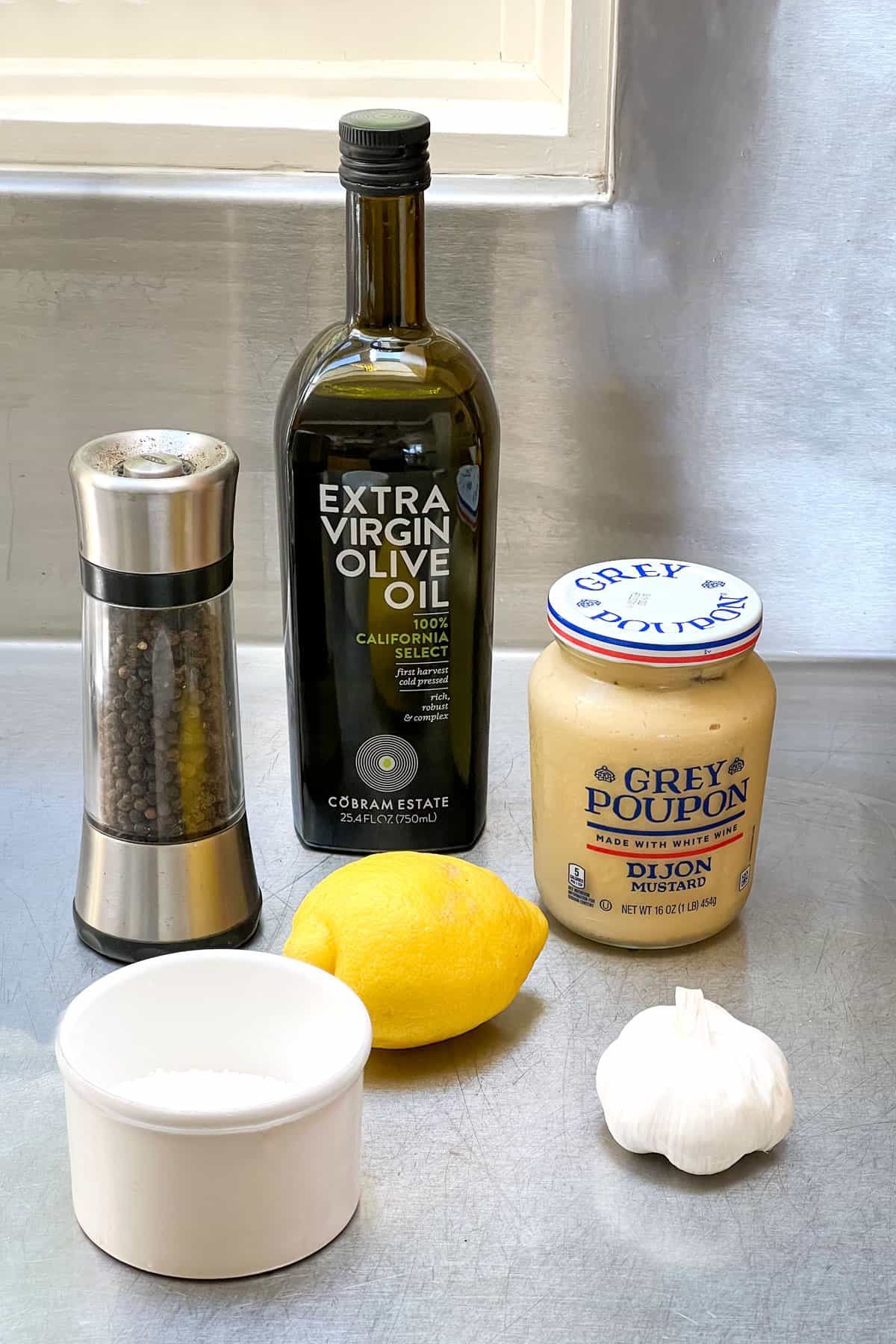 bottle of extra virgin olive oil, pepper grinder, jar of dijon mustard, a lemon, a white round salt cellar, and a bulb of garlic, all on a stainless steel countertop