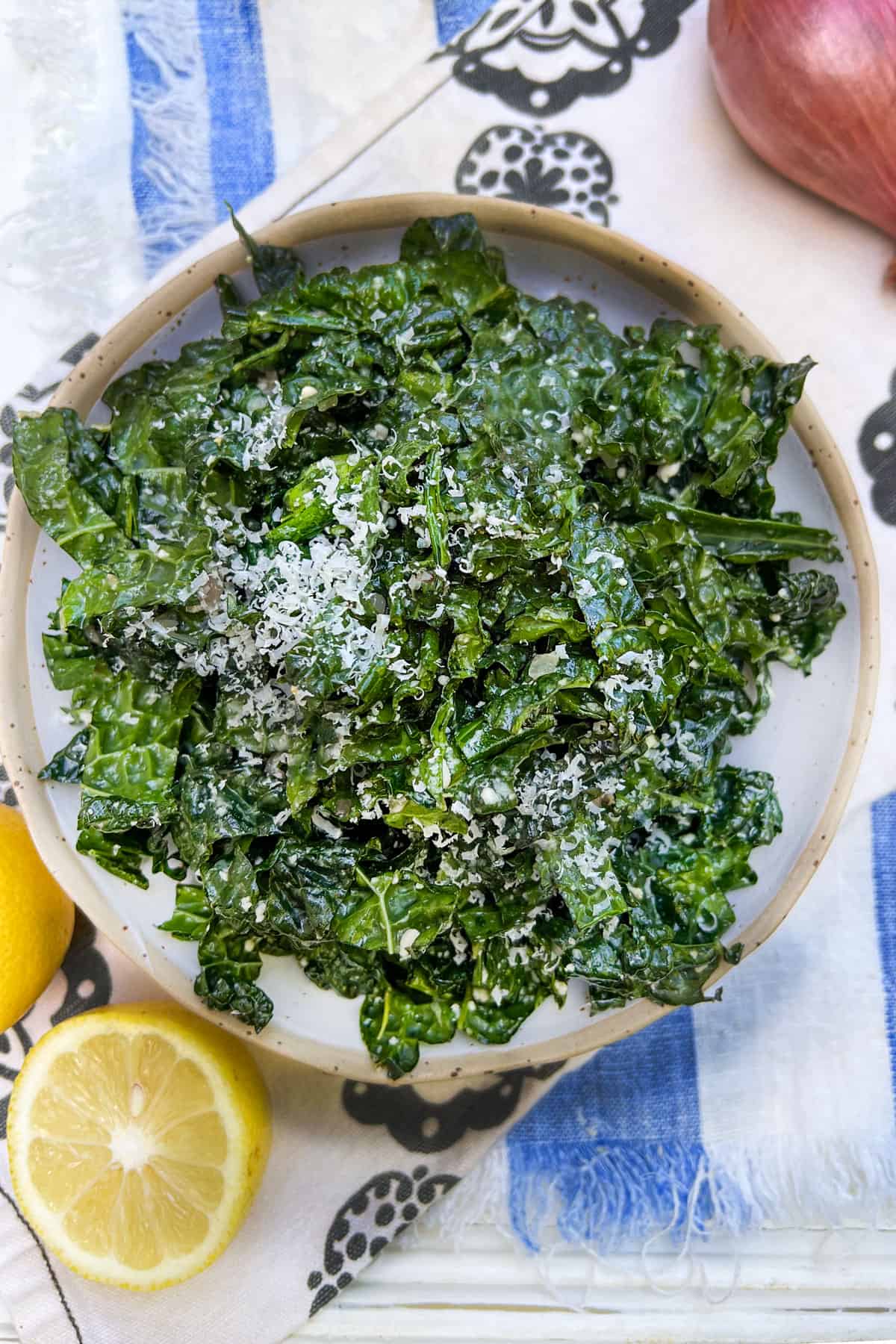 A bowl of shredded kale salad with dressing and a sprinkling of finely grated parmesan cheese. A cut lemon to one side and a shallot on the other side.