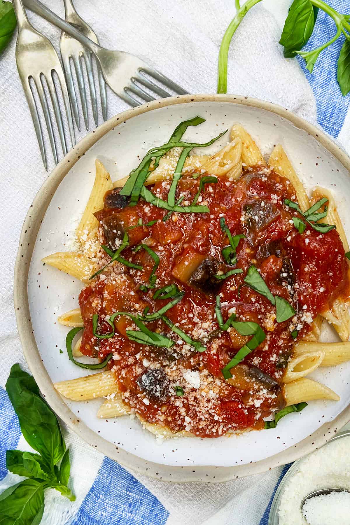 serving bowl filled with penne pasta topped with eggplant sauce, fresh slivered basil and basil leaves strewn around