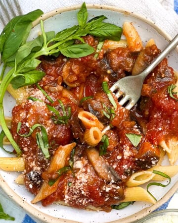 Bowl filled with penne pasta tossed with tomato eggplant sauce, fresh slivered basil and grated cheese, basil leaves strewn around