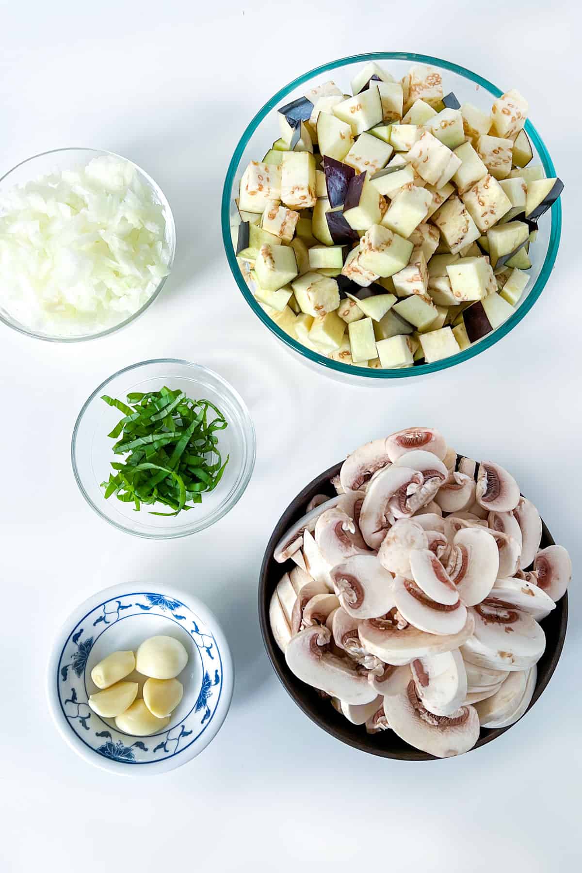 bowls of ingredients on a white countertop: sliced mushrooms, cubed eggplant, peeled garlic, chopped onions and slivered basil.