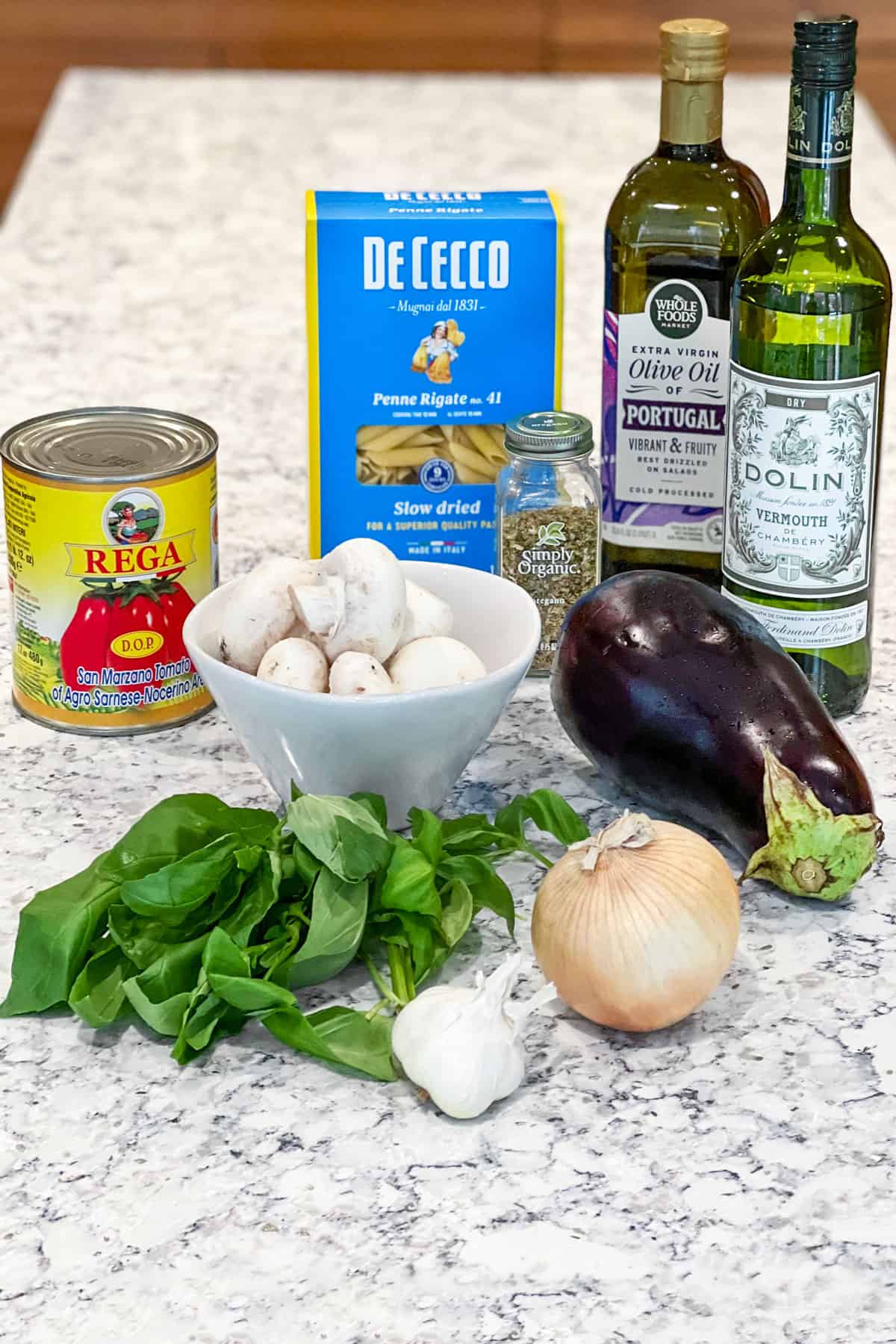 ingredients on a marble countertop: bottle of olive oil, bottle of dry vermouth, box of penne pasta, yellow onion, bulb of garlic, can of Italian tomatoes, bowl of white mushrooms, bunch of fresh basil, large eggplant.