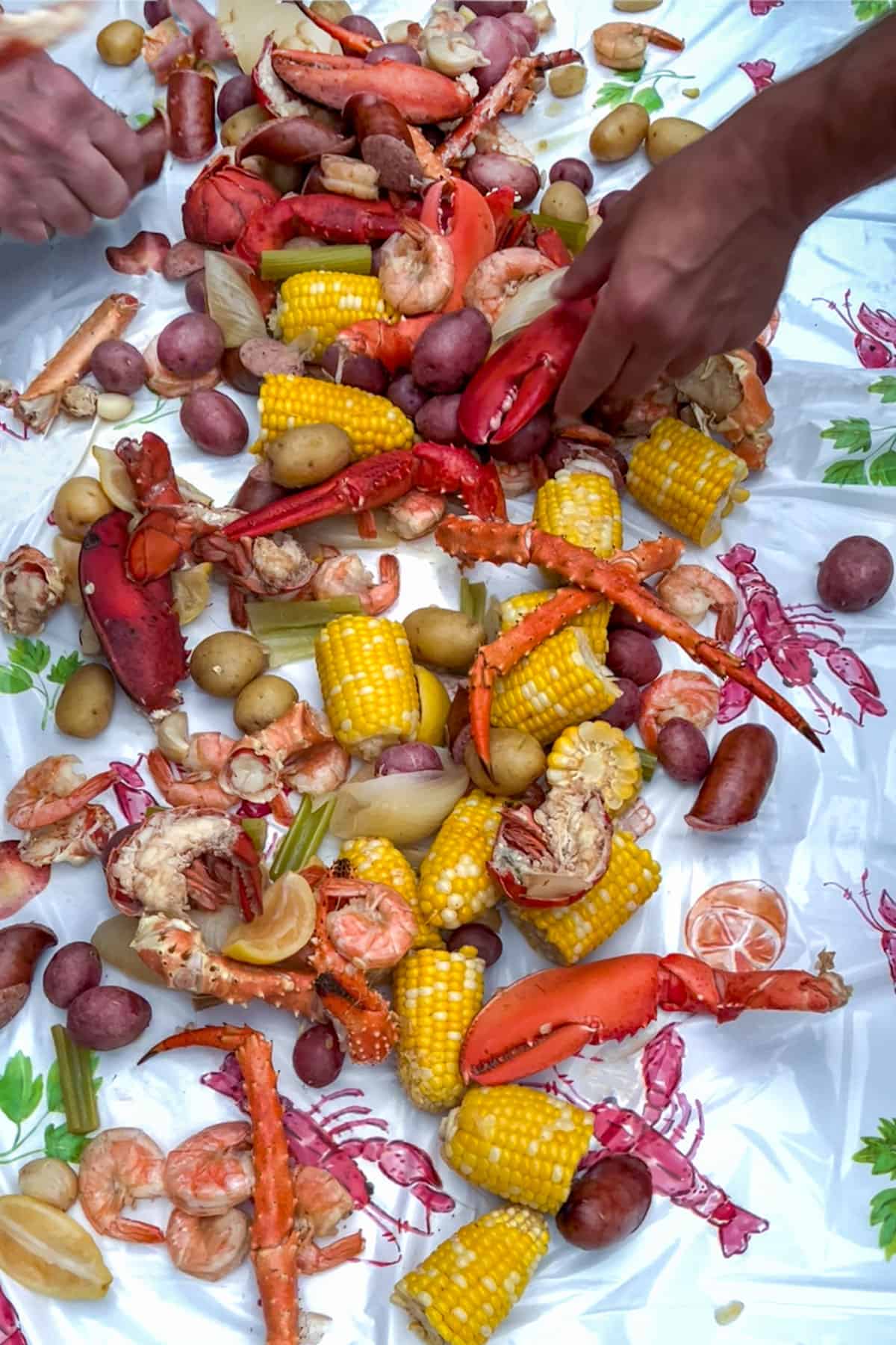 Hands reaching in to grab seafood from a table topped with a seafood boil that has lobster, crab legs, corn, potatoes, and shrimp