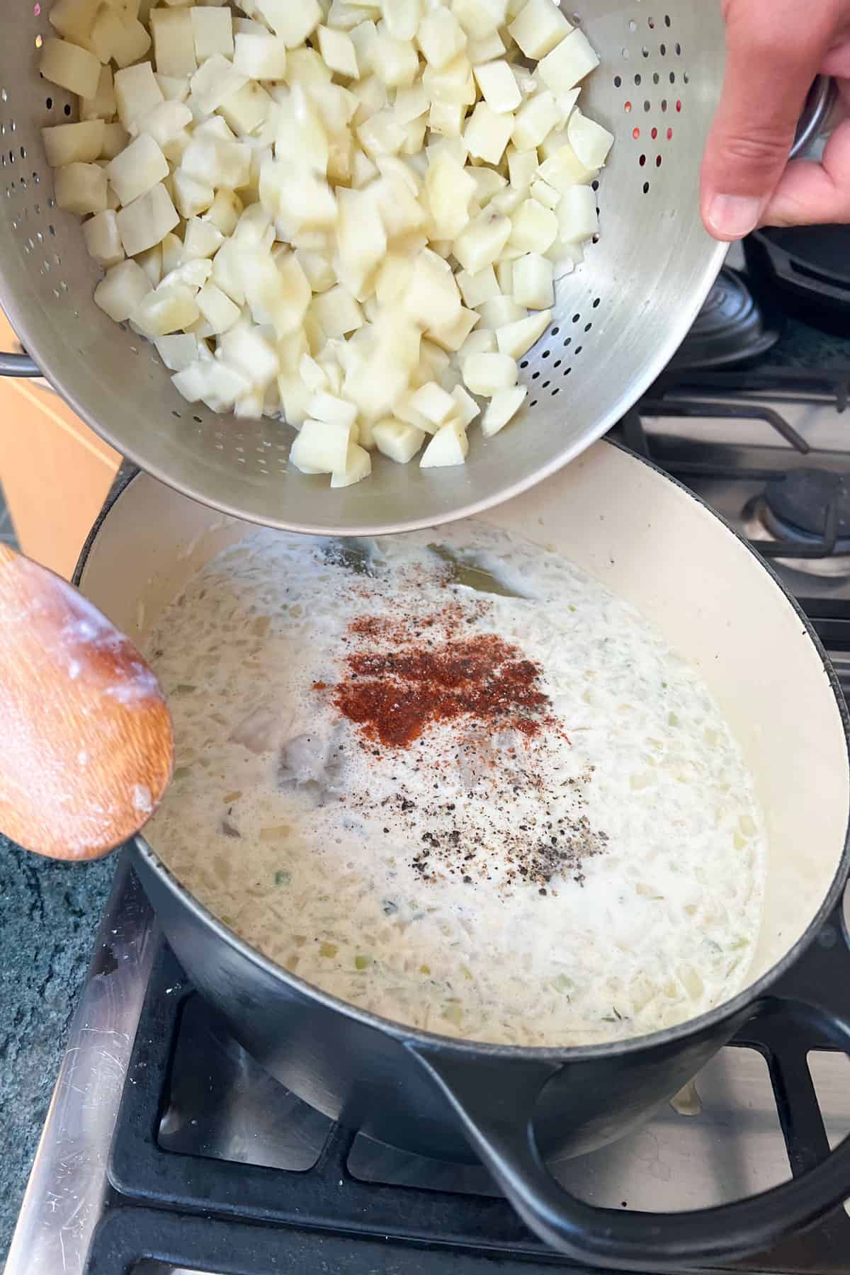 small cubes of partially cooked potatoes being poured from a steel colander into a dutch oven with shrimp chowder broth topped with pepper, salt and cayenne.