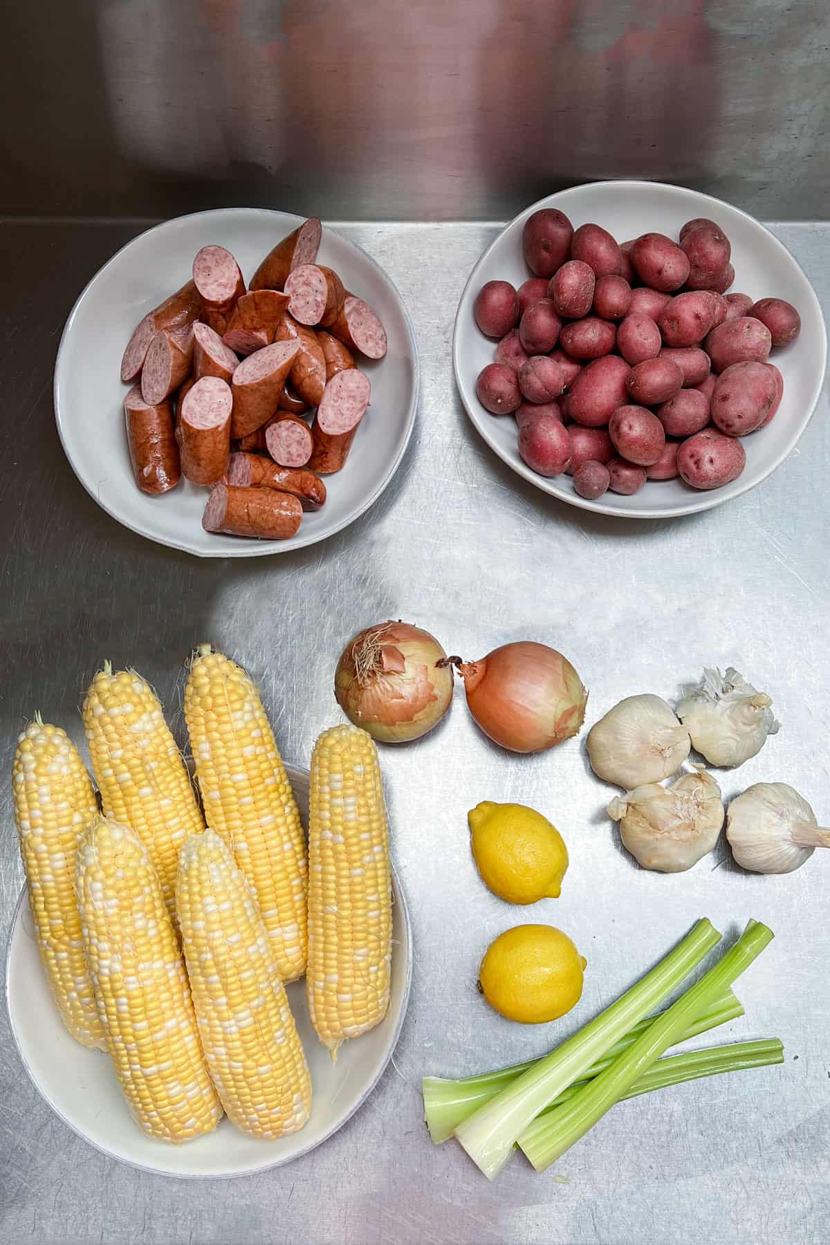 seafood boil ingredients on a stainless countertop: bowl of sliced sausages, bowl of small red potatoes, 6 ears of husked corn, two yellow onions, two lemons, 3 celery stalks and 4 garlic bulbs