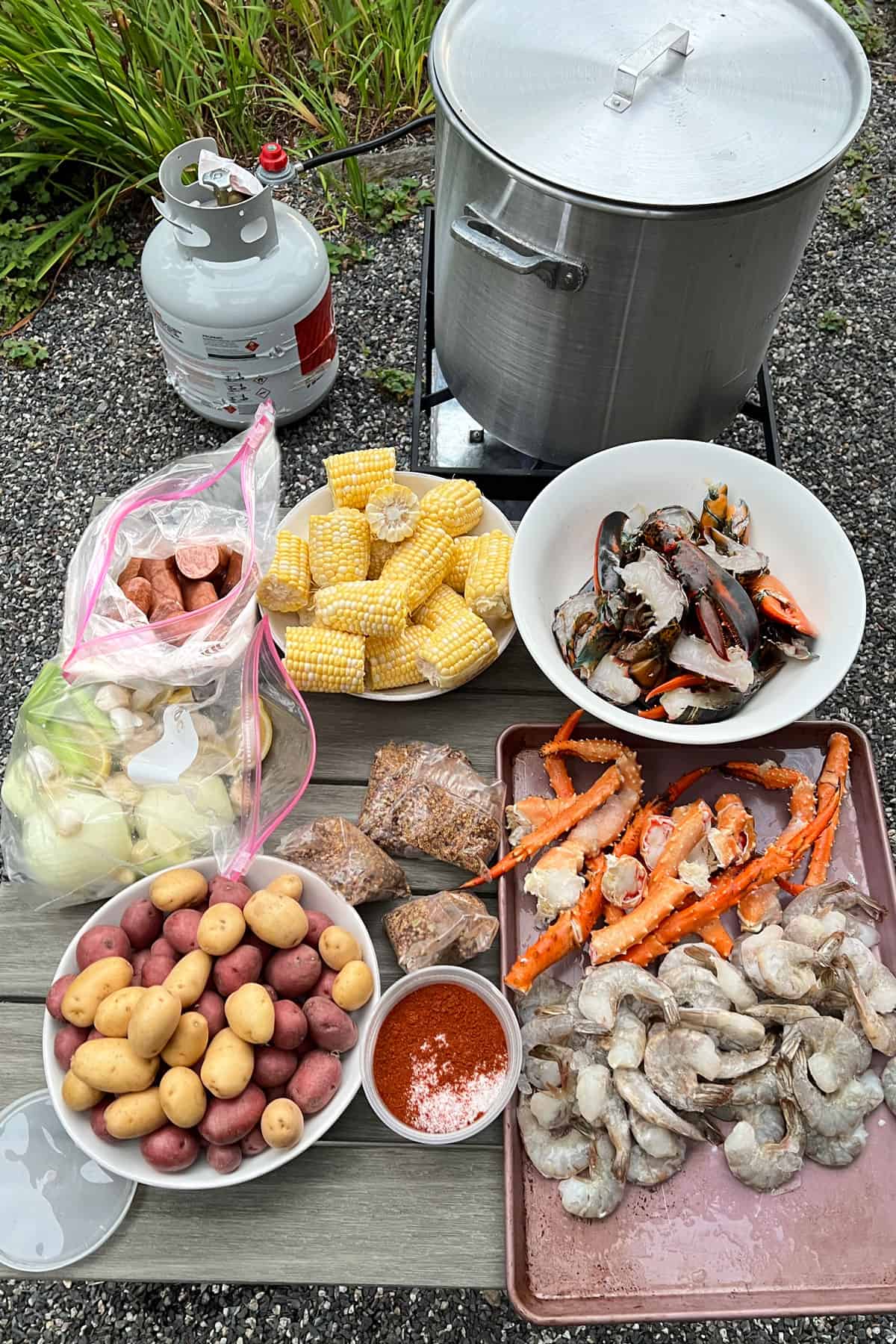 Seafood boil ingredients on an outdoor table near a big boiling pot with a propane tank. Bowl of lobster claws and tails, bowl of small potatoes, tray of shrimp and crab legs, bowl of corn, bag of sausages, bag of onions, celery and garlic, bags of spices and a container of salt and cayenne.