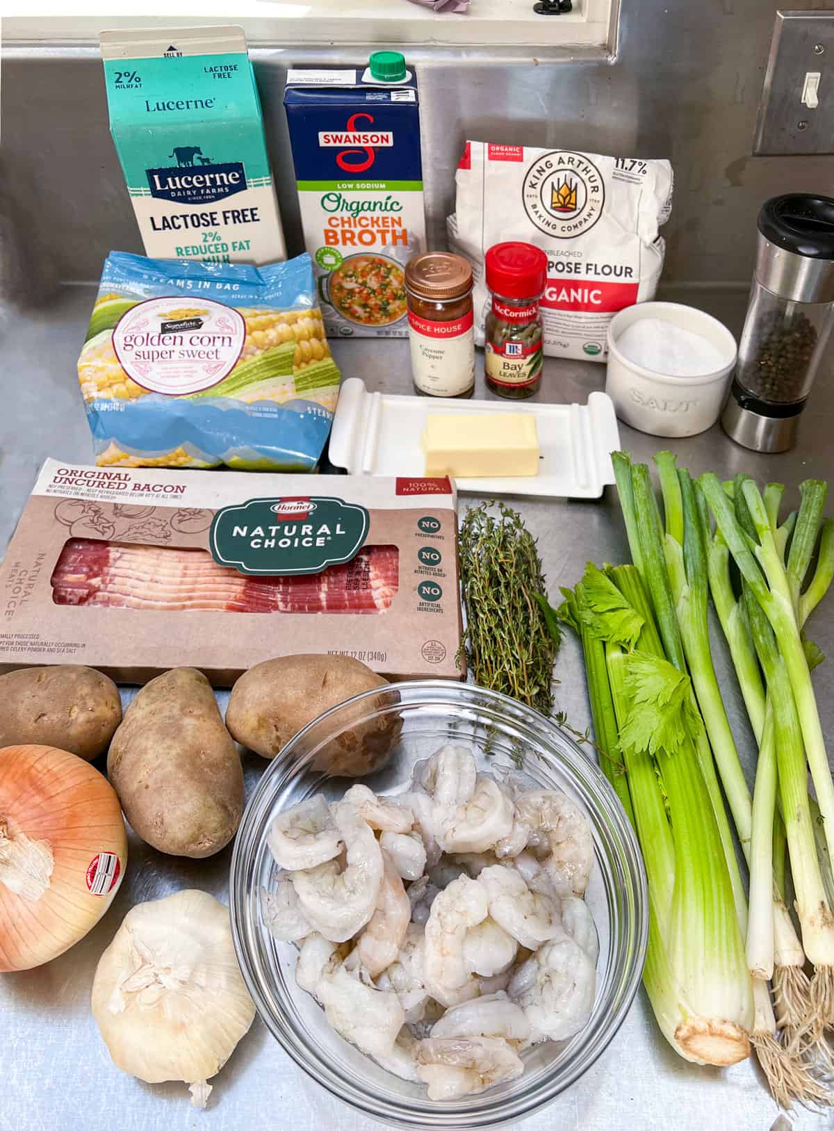 Ingredients on a stainless steel counter: bowl of raw shrimp, bunch of celery, bunch of scallions, 3 russet potatoes, an onion, a bulb of garlic, a pack of bacon, some fresh thyme sprigs, a pack of frozen corn, a box of chicken broth, box of milk, bag of four, butter, salt cellar, pepper grinder, bottle of bay leaves, bottle of cayenne pepper.