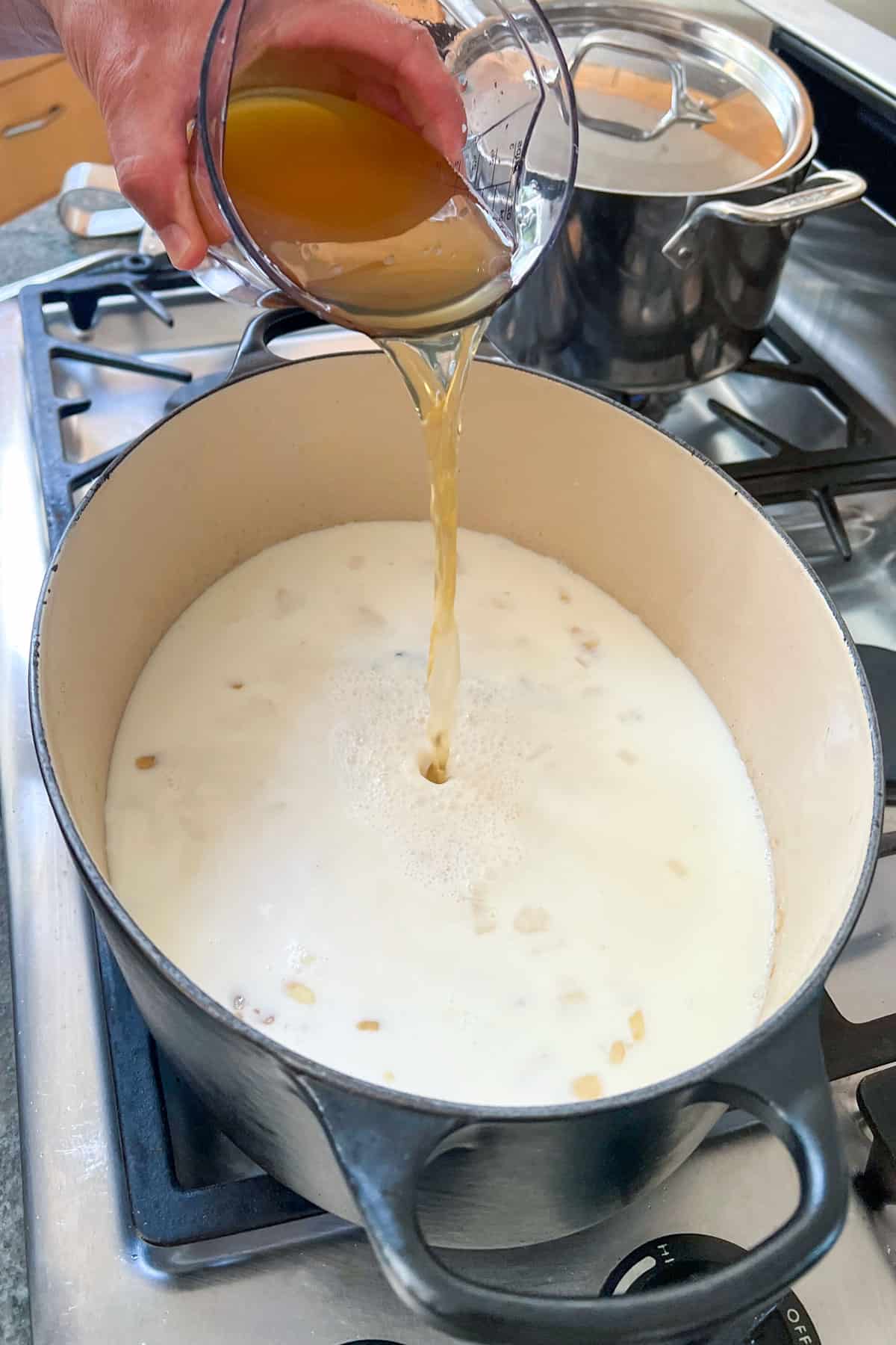 chicken broth being poured from above, into a dutch oven filled with a milky broth.