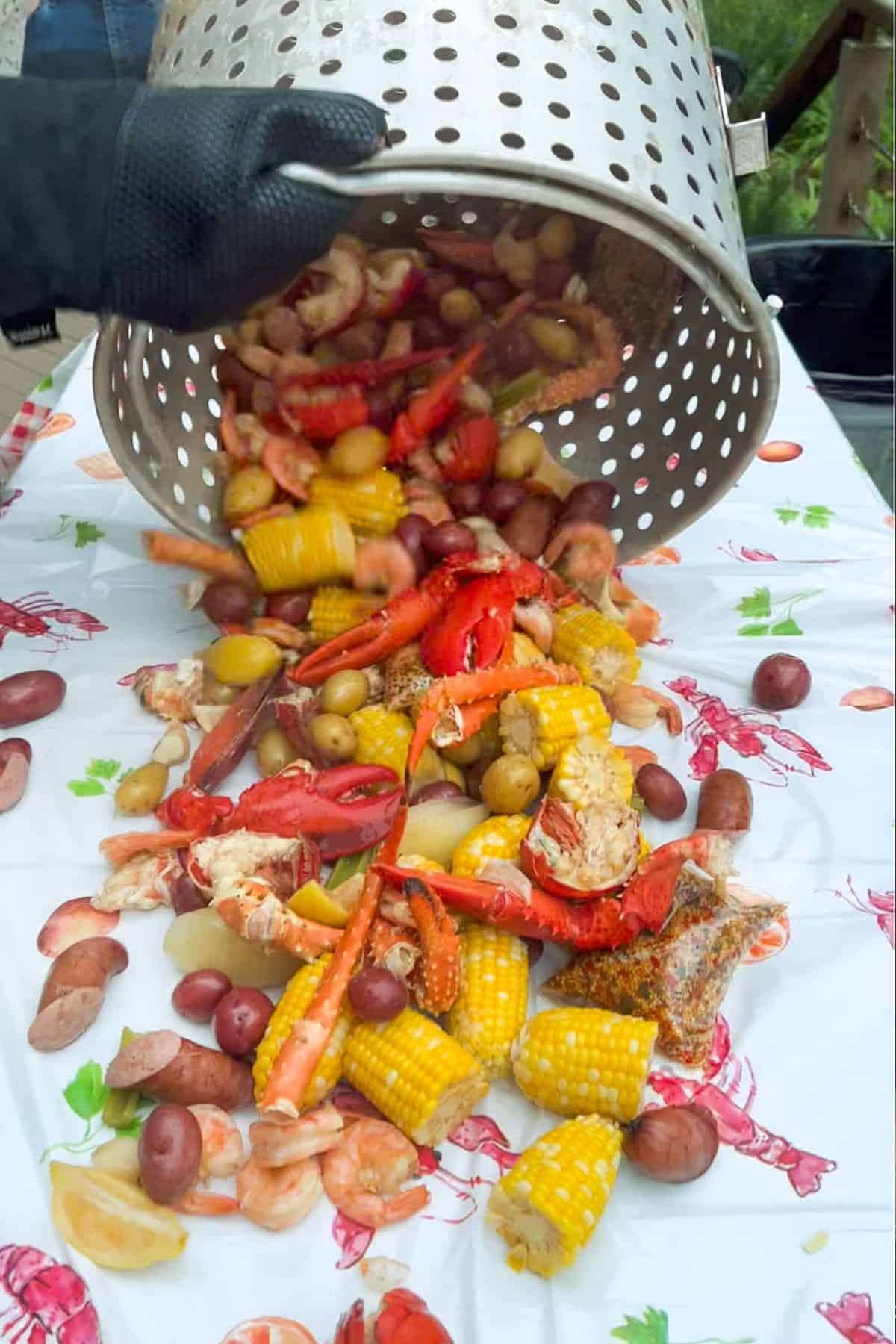 the contents of a huge seafood boiling pot strainer being poured onto a large table. Corn, lobster tails and claws, potatoes, clams, crab legs and sausages.