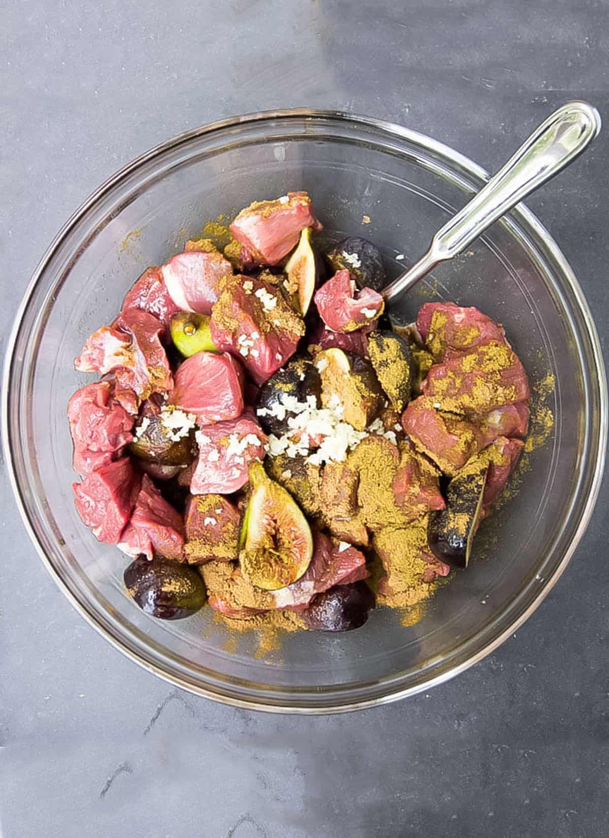Glass mixing bowl filled with cubes of lamb, fresh figs, cumin and garlic. 