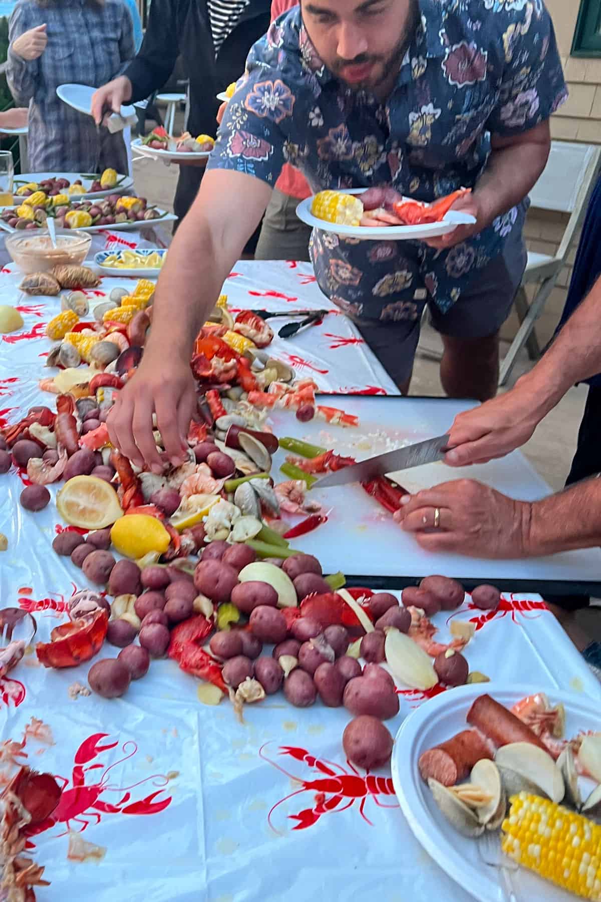 People standing around a table covered with seafood, corn and potatoes, reaching out to grab stuff to put on their paper plates.
