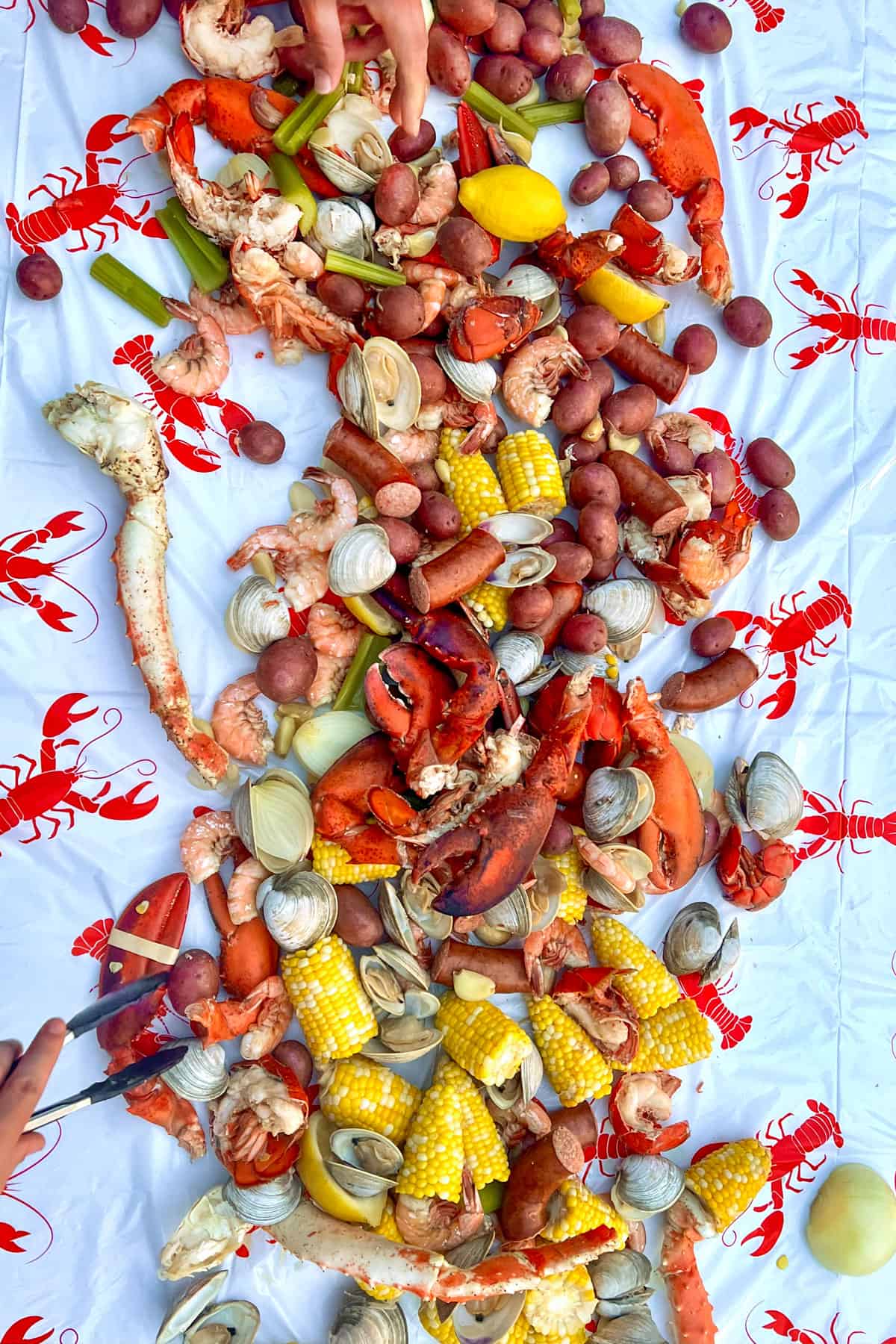 Lots of cooked lobster claws and tails, corn, lemons, clams, crab legs, baby red potatoes, celery, clams and sausages spread out on a table covered with a lobster-printed plastic table cloth.