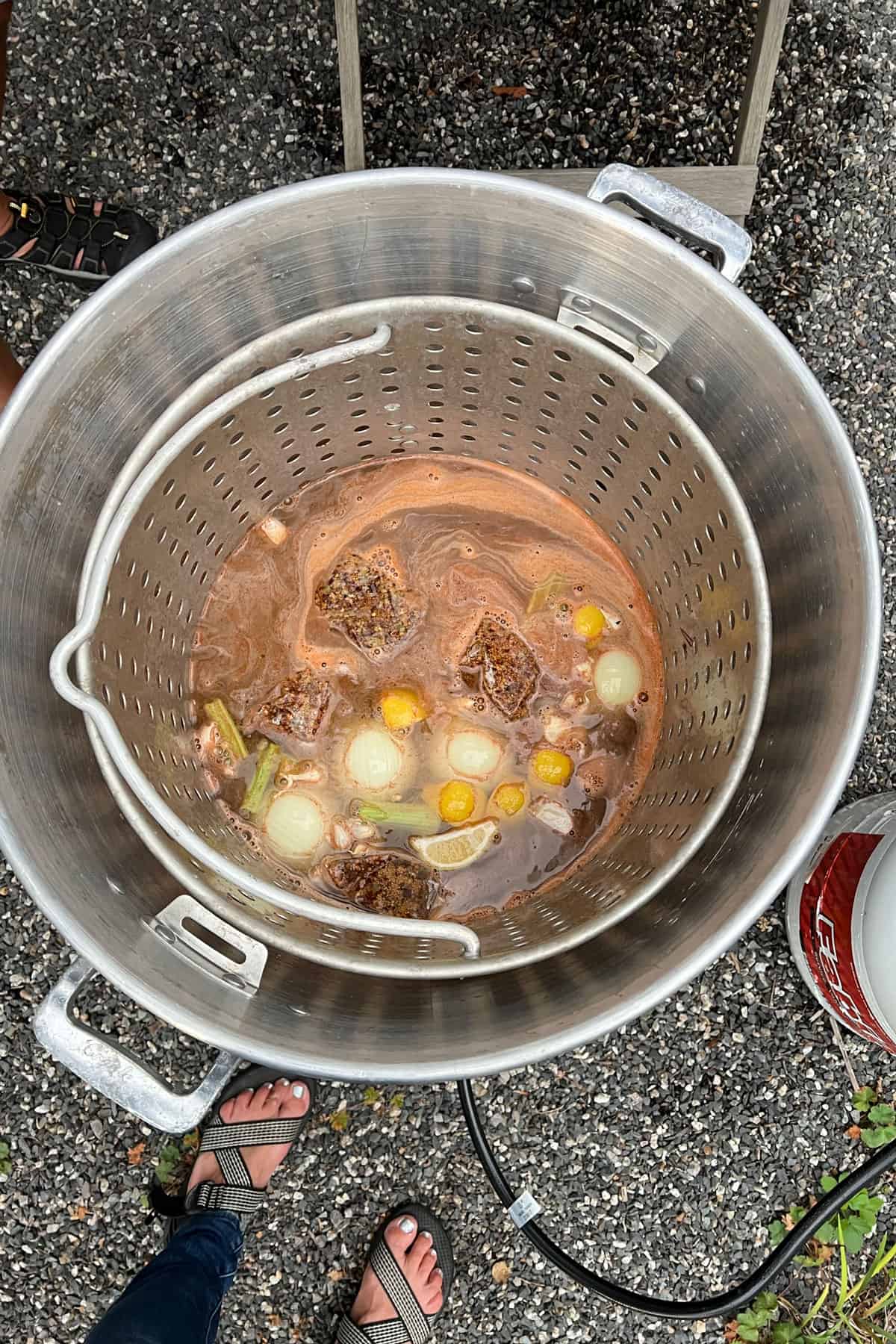 looking down into an outdoor seafood boiling pot with strainer, filled with water, spices, onions, lemons, celery and garlic