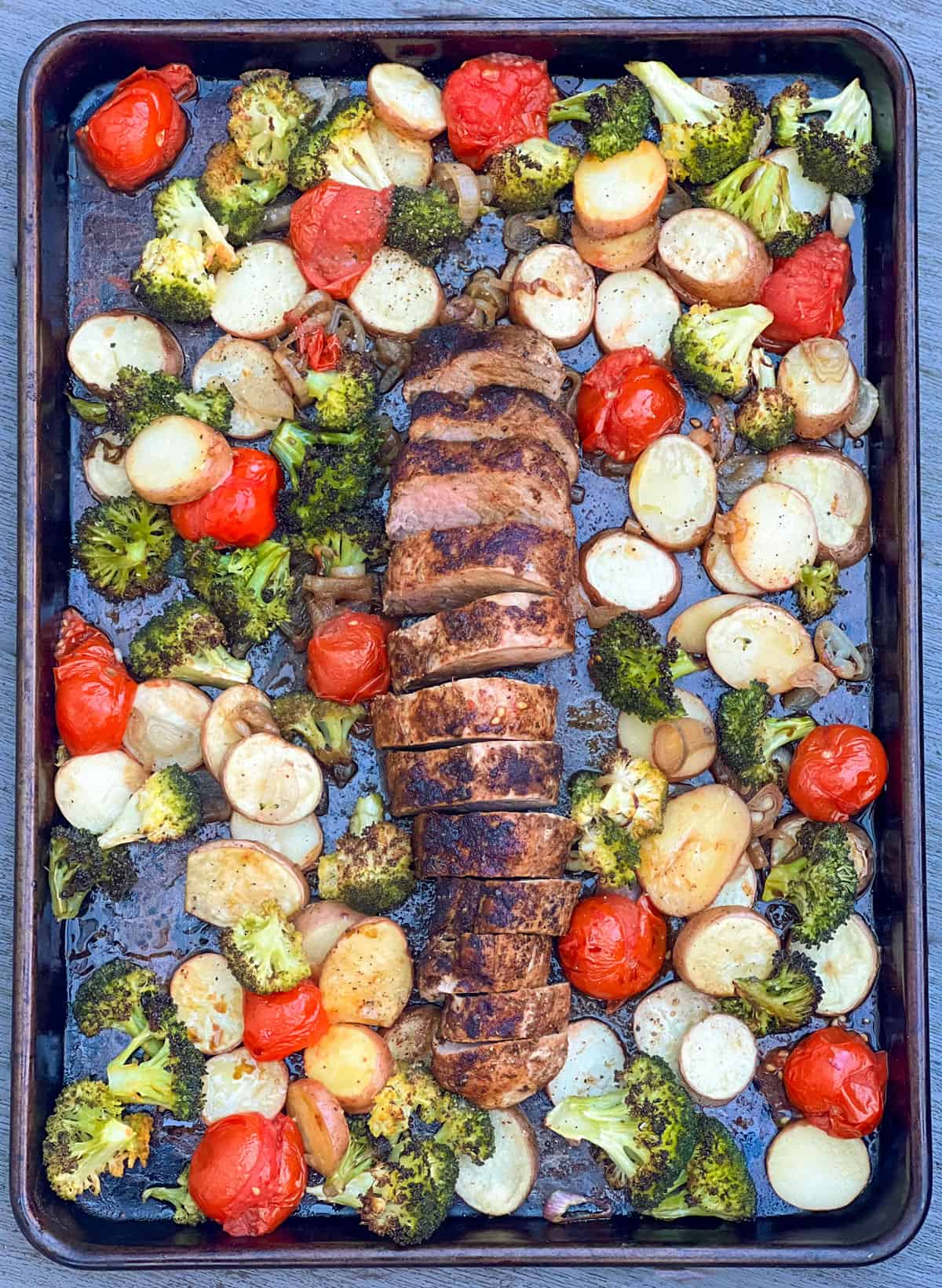 Roasted sliced pork tenderloin on a sheet pan surrounded by roasted potatoes, broccoli and cherry tomatoes.