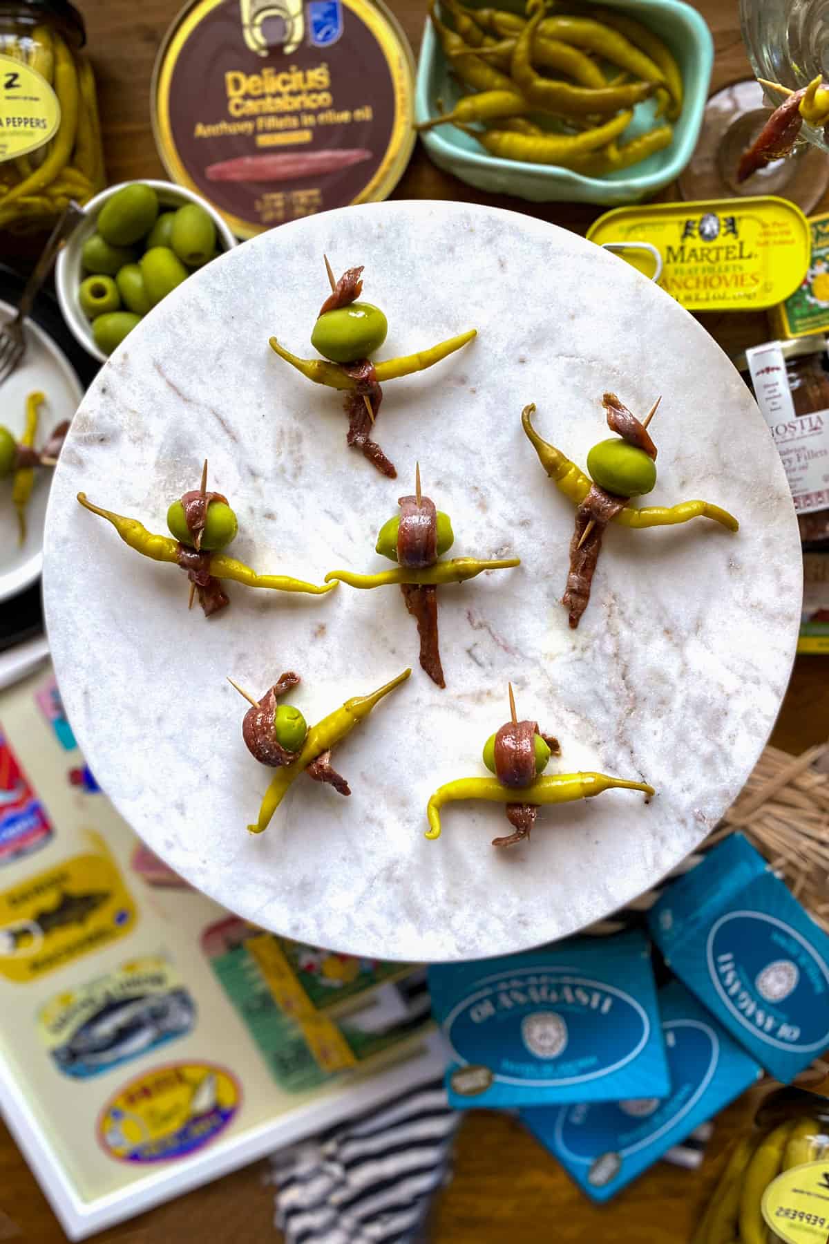 Six anchovy, olive and guindilla pepper skewers on a marble cake stand with a background of tinned seafood