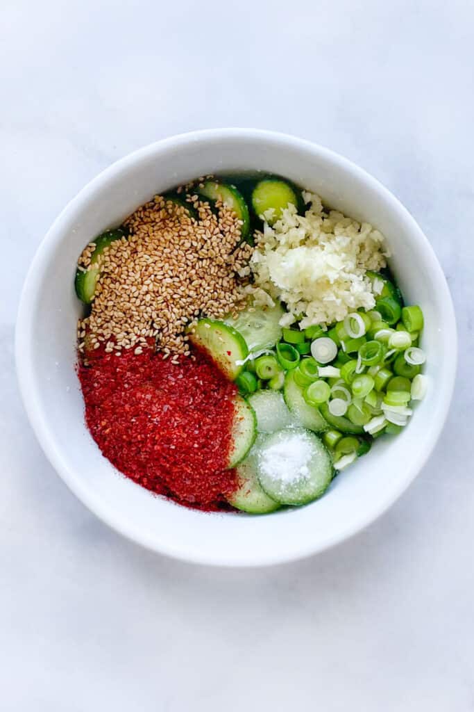 cucumber slices in a bowl with gochugaru, sesame seeds, sugar, garlic and scallions on top