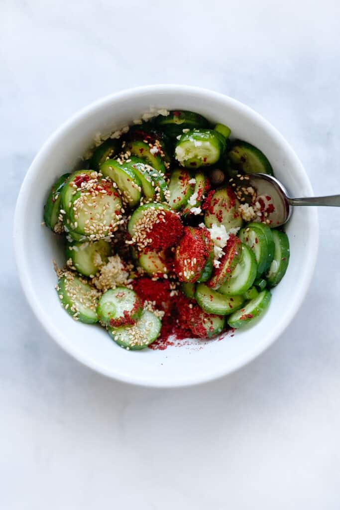 cucumbers in a bowl being mixed with gochugaru, sesame seeds, sugar, garlic and scallions on top