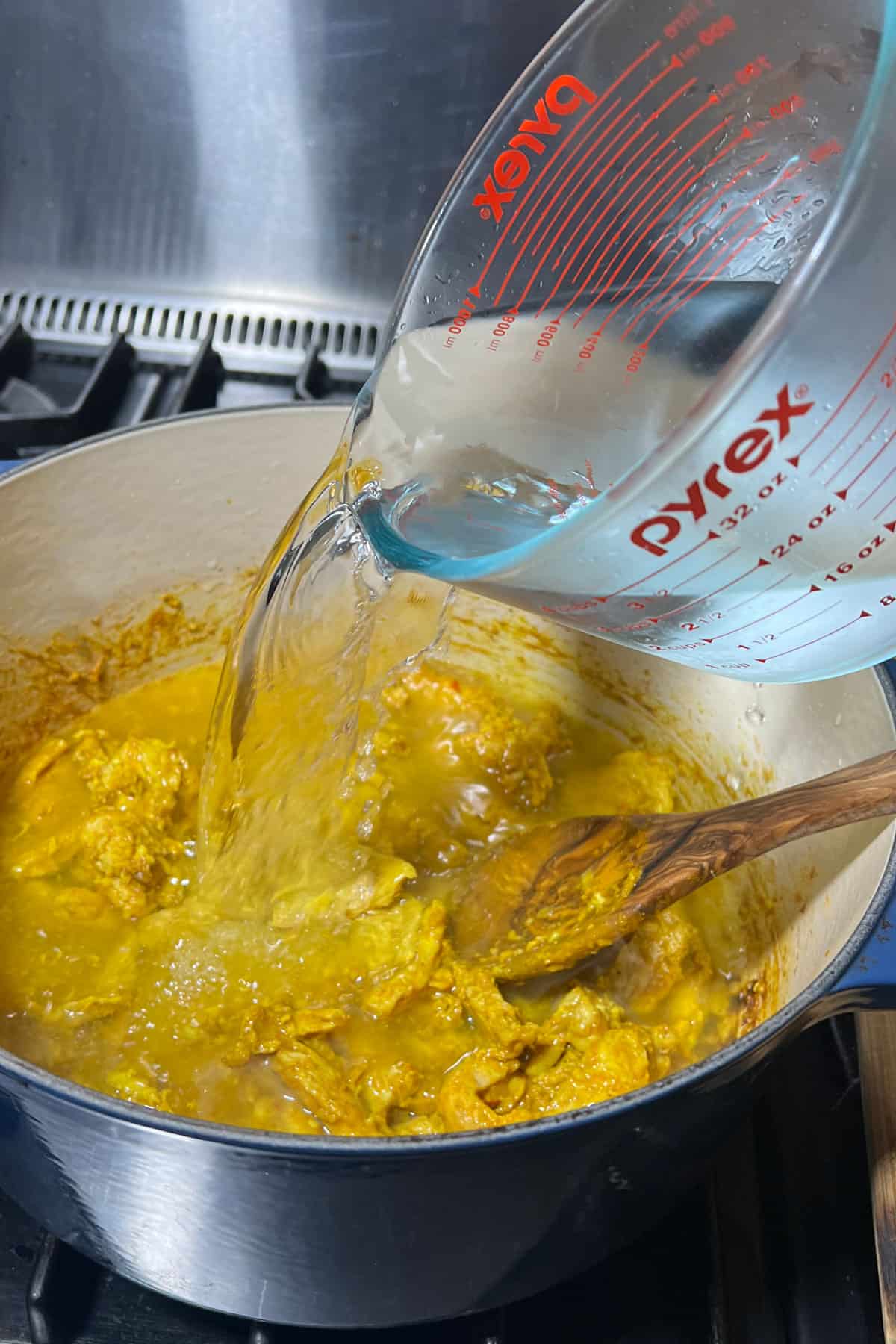 Water being poured into a Dutch oven filled with sauteed turmeric chicken.