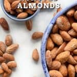 Salted roasted almonds in a big bowl and small bowl with almonds scattered next to them