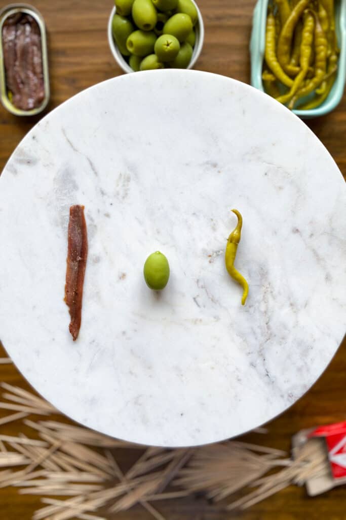 an anchovy, a guindilla pepper and a frescatrano olive sitting on a marble cake stand.