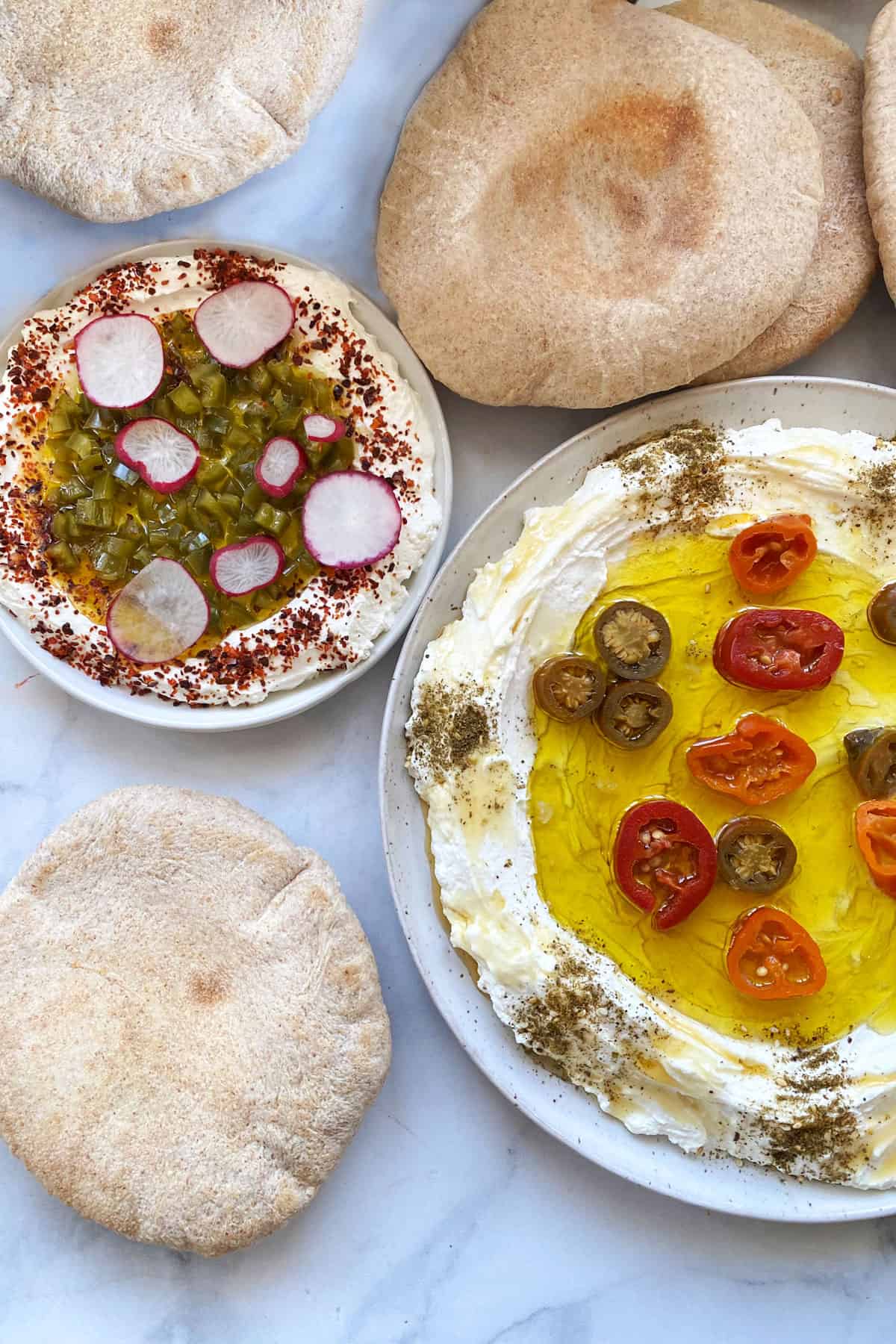 two plates of labneh with olive oil and pickled jalapenos and pita in background