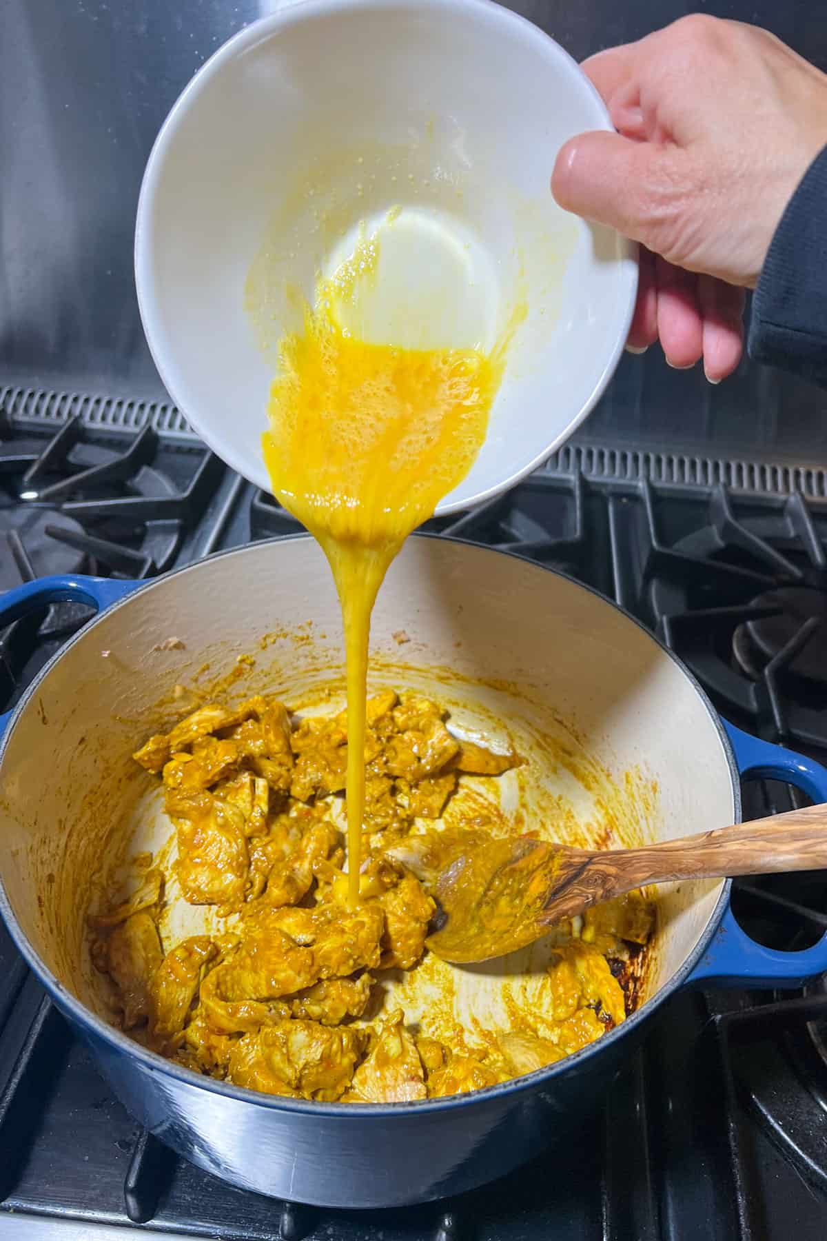 pot of yellow colored sauteed chicken and a small bowl above pouring a whisked egg into the pot.