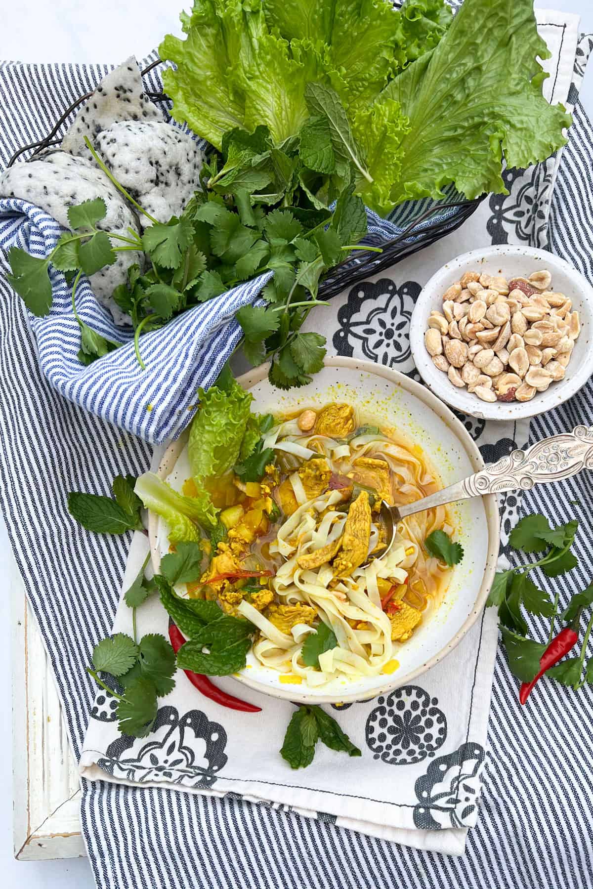 basket of herbs, lettuces and puffy sesame rice crackers next to a bowl of noodles and chicken in turmeric broth and a small bowl of peanuts