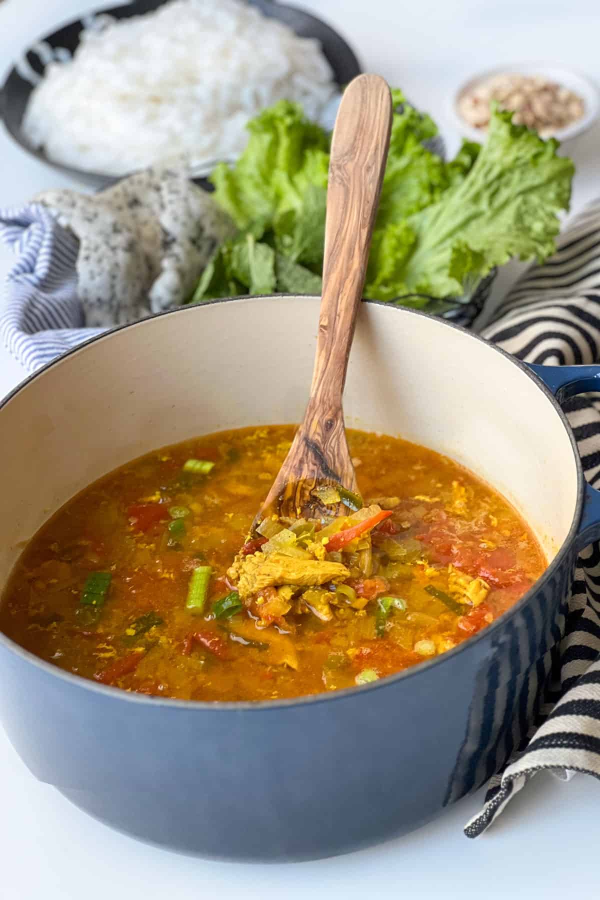 Round Dutch oven half filled with a pieces of chicken in broth with bits of tomato and scallion, lettuce and puffy rice crackers in the background