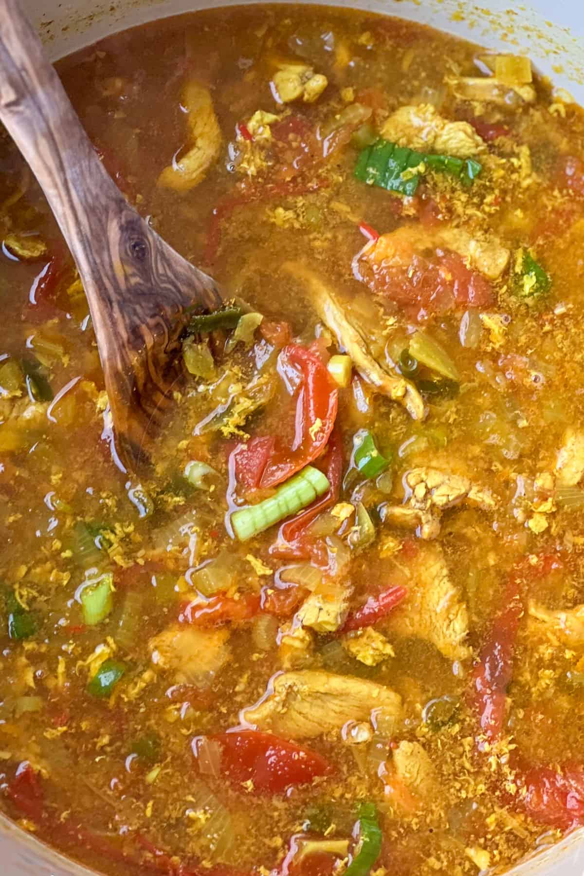 Turmeric broth with chicken, tomatoes, chopped scallions and a wooden spoon in the liquid.