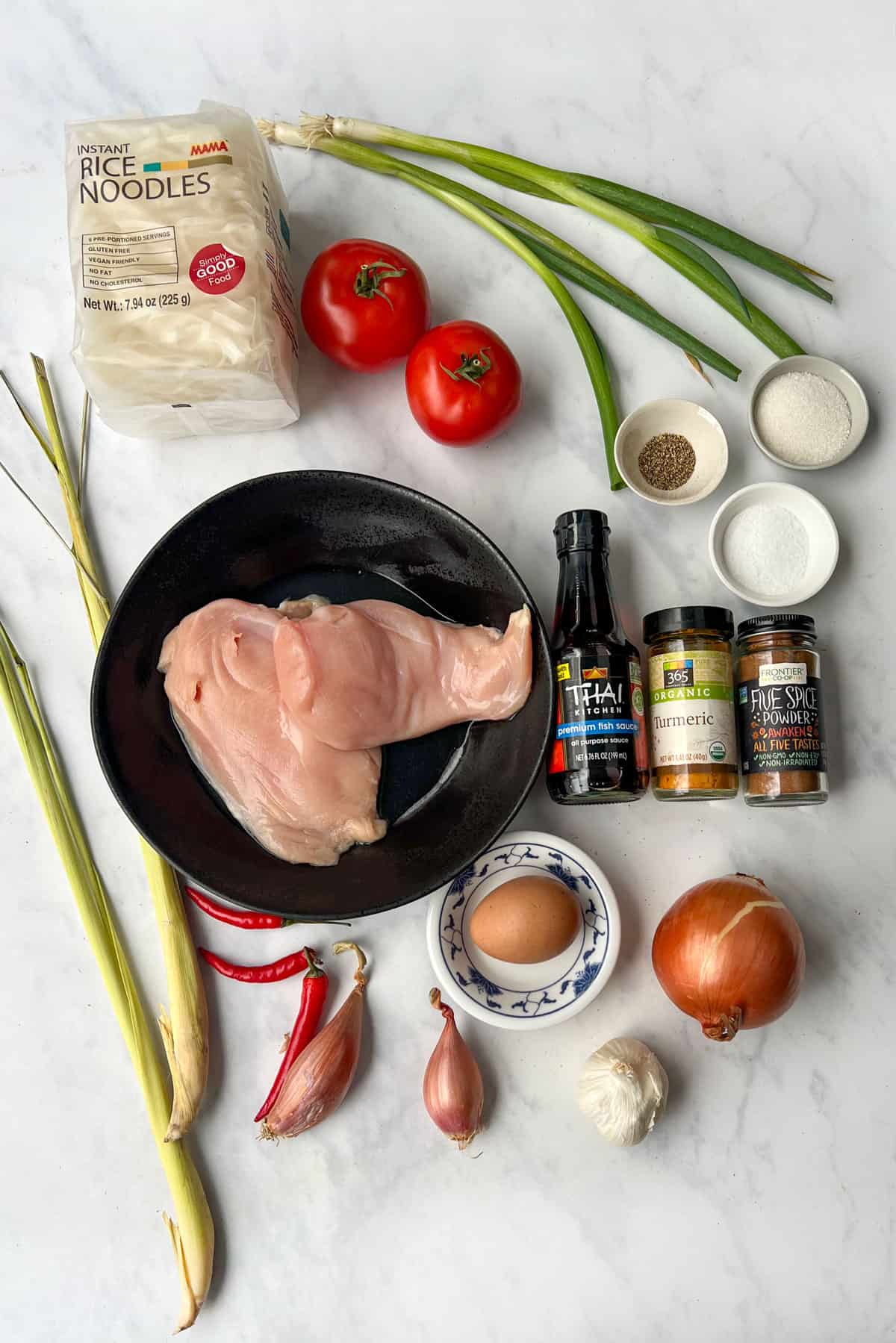 ingredients on a marble countertop: two boneless chicken breasts, two stalks lemongrass, two tomatoes, two scallions, a yellow onion, a shallot, an egg, a garlic bulb, a pack of rice noodles, bottle of fish sauce, jar of turmeric, gar of 5 spice powder.