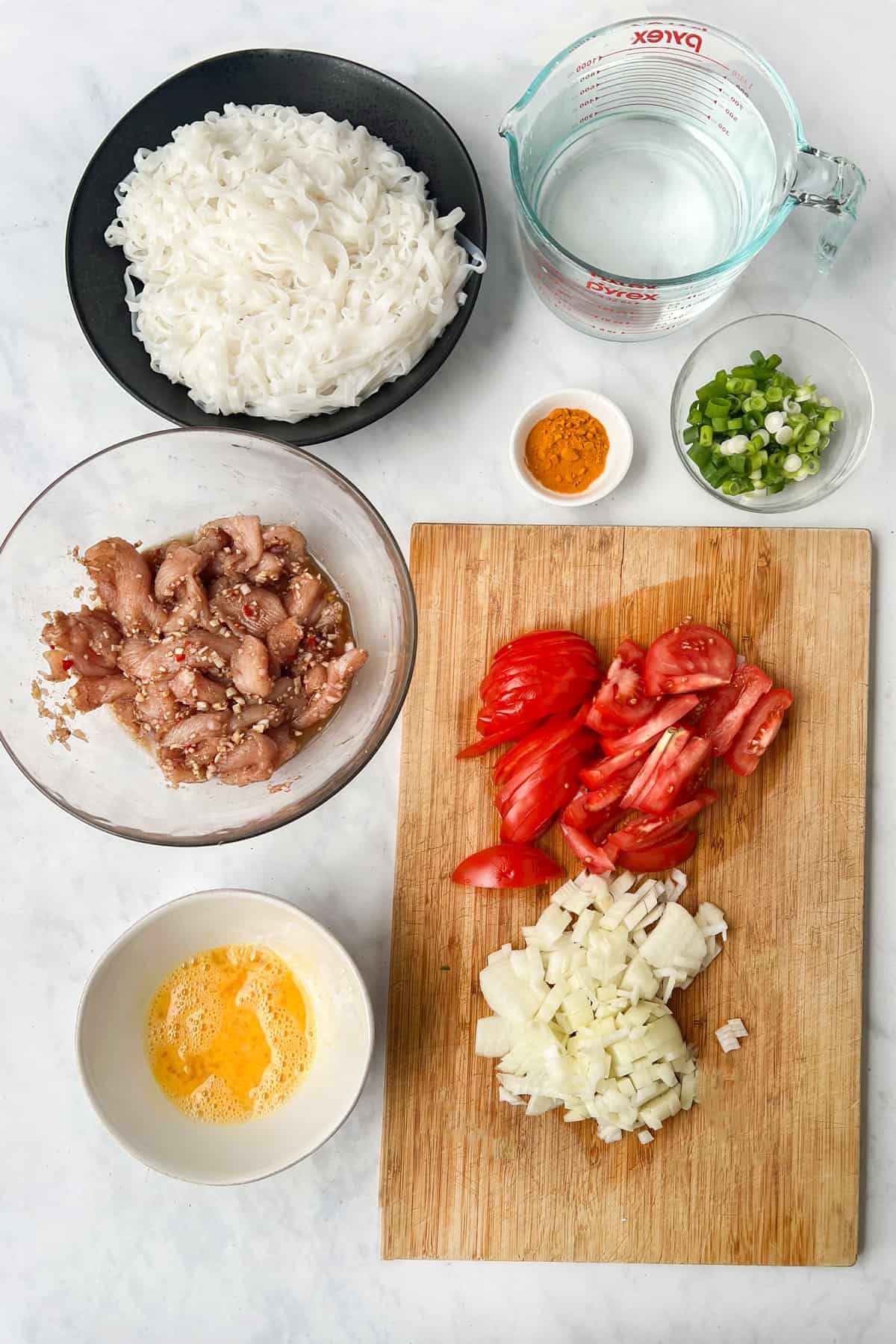 Ingredients on a marble countertop. Chopped onions, sliced tomatoes, chopped scallions, sliced chicken breast, a whisked egg, turmeric in a small bowl, a 4-cup measuring cup filled with water.