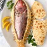 a full skin-on snapper inside a cut-open salt pastry with the top of the pastry lying next to it on a plate garnished with parsley and lemon