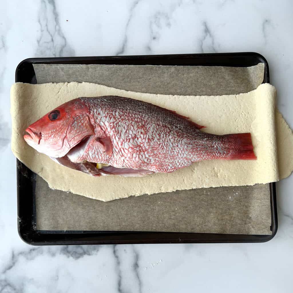 whole red snapper stuffed with lemon slices atop rolled out salt dough on a parchment-lined baking tray