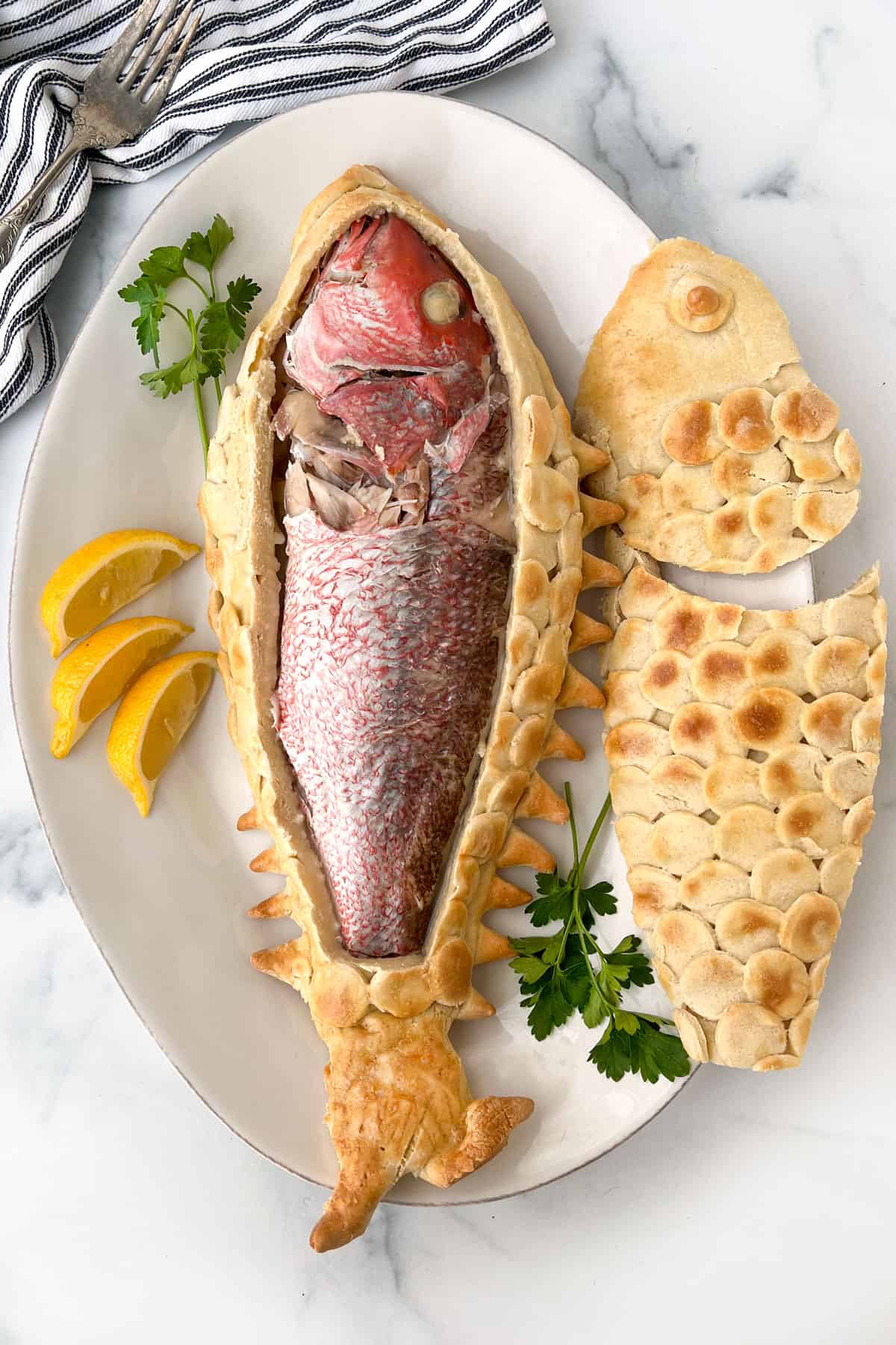 a full skin-on snapper inside a cut-open salt pastry with the top of the pastry lying next to it on a plate garnished with parsley and lemon