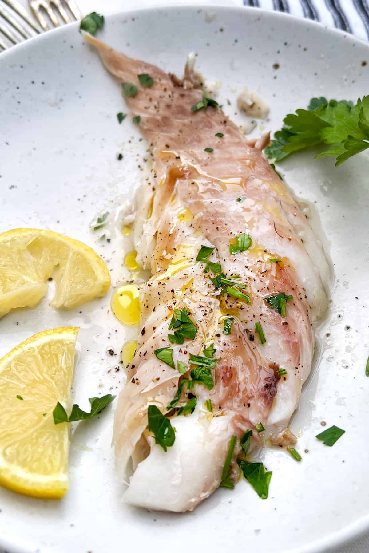 A portion of red snapper on a plate with olive oil, lemon slices and parsley