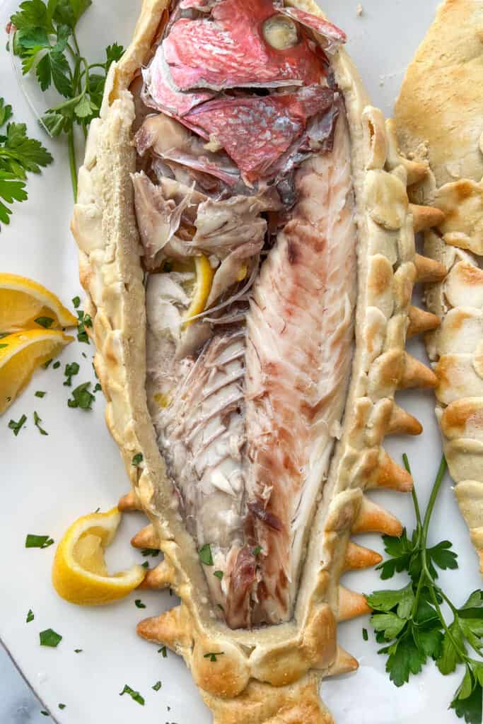 A fully cooked whole red snapper exposed from it's salt crust and partially eaten with lemon slices and parsley next to it