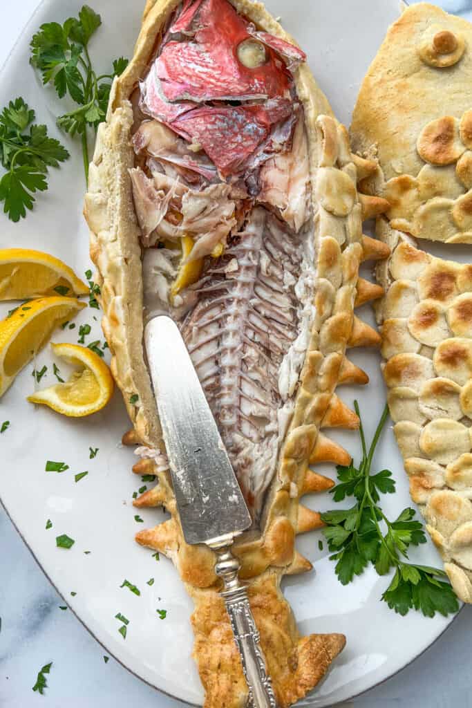 A fully cooked whole red snapper exposed from it's salt crust and half eaten with lemon slices and parsley next to it and a knife