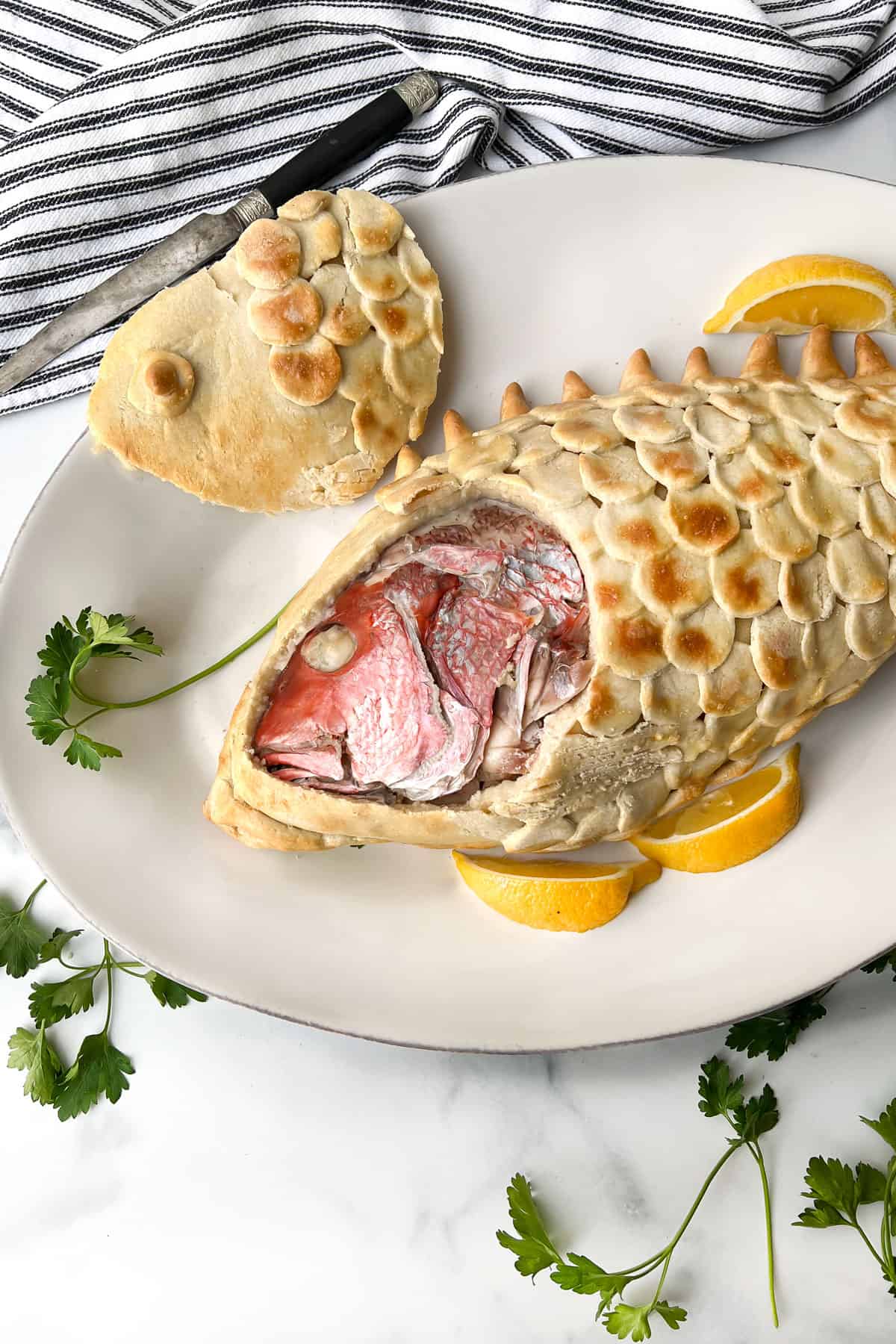 A baked fish in salt pastry with the pastry cut so the fish head is exposed   lying on a plate garnished with parsley and lemon with a plate of lemon wedges in the background