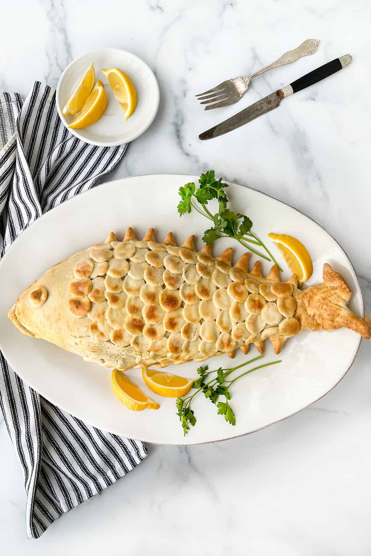Baked fish in salt pastry shaped like a fish with scales on top of a plate with lemons and parsley and a knife and fork in the background