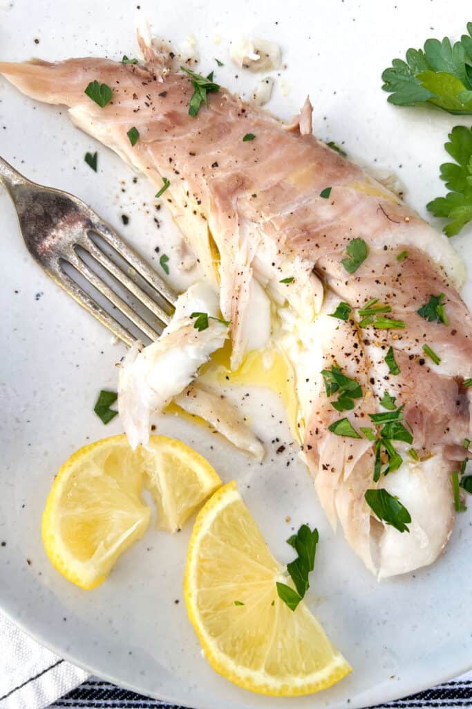 A portion of red snapper on a plate with olive oil, lemon slices and parsley next to a fork