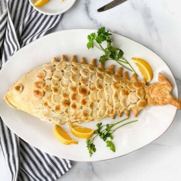 Salt pastry shaped like a fish with scales on top of a plate with lemons and parsley and a knife and fork in the background