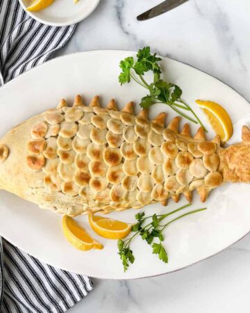 Salt pastry shaped like a fish with scales on top of a plate with lemons and parsley and a knife and fork in the background