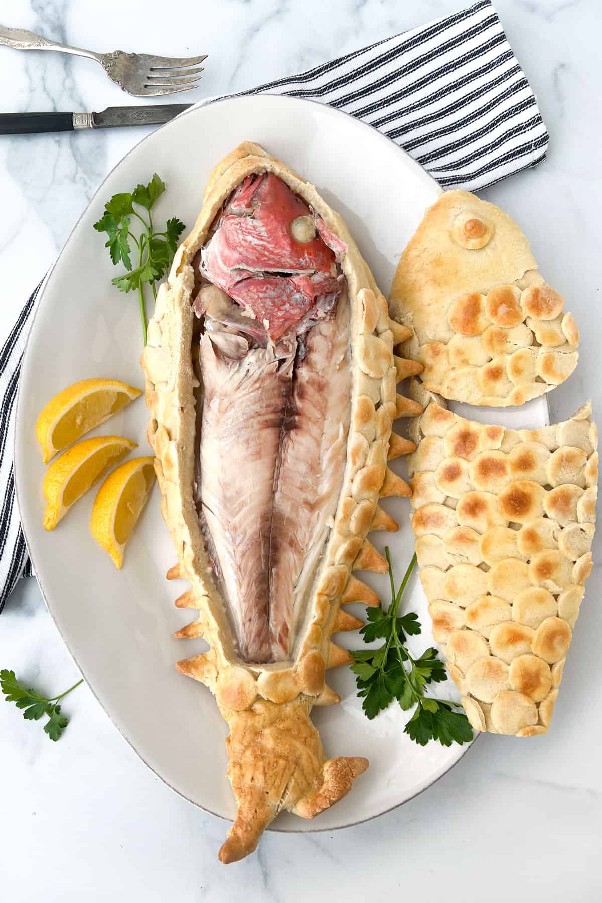 a full snapper inside a cut-open salt pastry with the top of the pastry lying next to it on a plate garnished with parsley and lemon