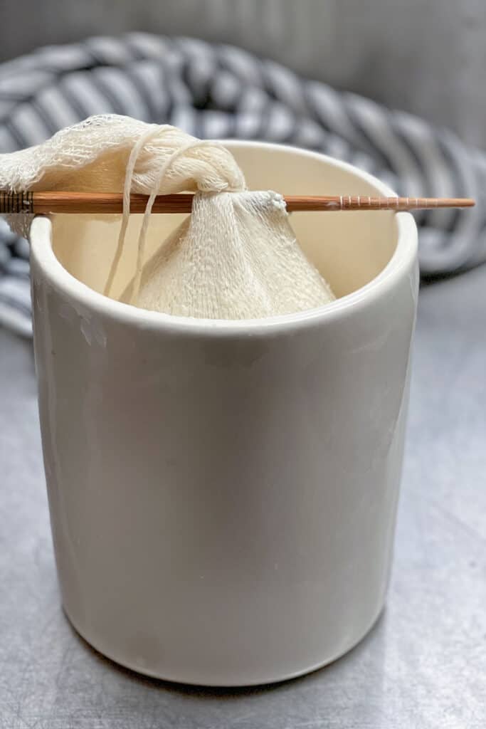 salted greek yogurt in cheesecloth skewered by a chopstick and hanging inside a jar