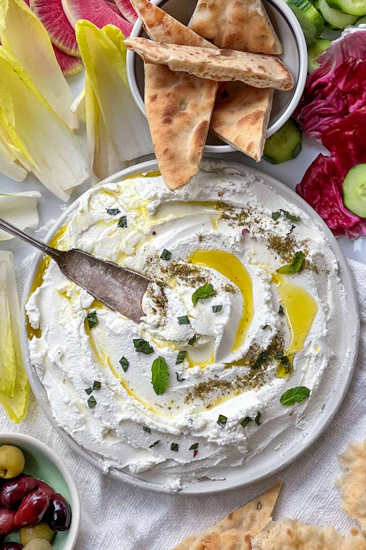 labneh dip on a plate with olive oil and mint and a knife surrounded by vegetables and pita