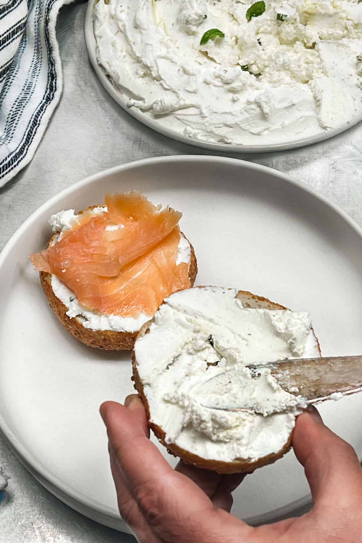 a bagel being smeared with labneh with one half covered in smoked salmon and a plate of labneh in the background