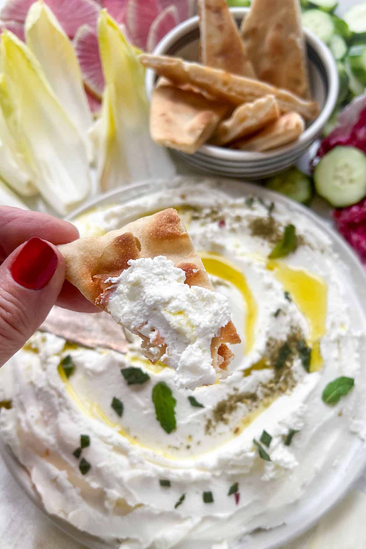 pita being dipped into labneh on a plate surrounded by vegetables and pita