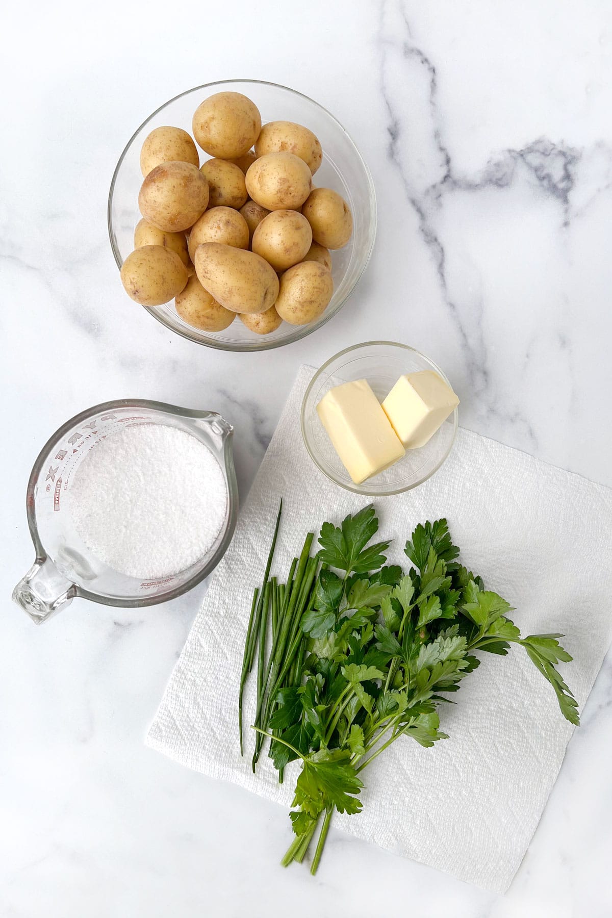 golden new potatoes, half a stick of butter divided in half, a cup of salt and some parsley and chives sitting on marble