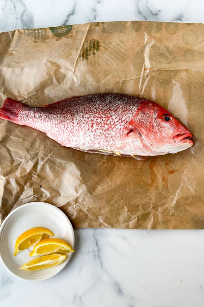 A whole red snapper lying on parchment paper with a small plate with lemon slices next to it